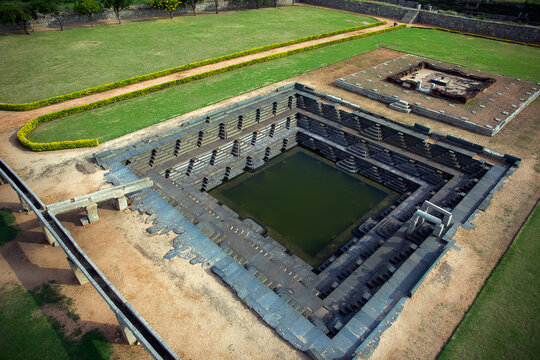 Aerial view of the stepped square Pushkarani tank, its dark waters contrasting with the pale stone steps and surrounding green lawns, Hampi, Karnataka, India.