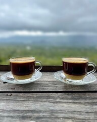 Two glass cups of coffee on a wooden table with a green mountain view
