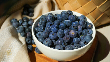 A bowl of ripe blueberries sits on a wooden board with a woven basket in the background, creating a rustic and natural setting ideal for showcasing healthy, fresh, and organic food. Close up