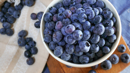A bowl of ripe blueberries sits on a wooden board with a woven basket in the background, creating a rustic and natural setting ideal for showcasing healthy, fresh, and organic food. Close up