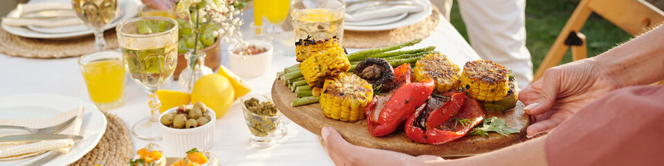 Caucasian woman serving grilled vegetables including corn, asparagus, mushrooms, and red peppers on wooden platter during outdoor meal with people sitting at table with drinks and food