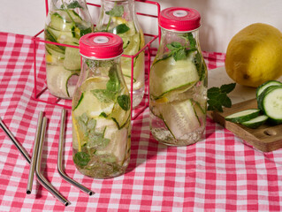 Glass bottles with red lids, filled with detox water containing cucumber and lemon slices and mint...