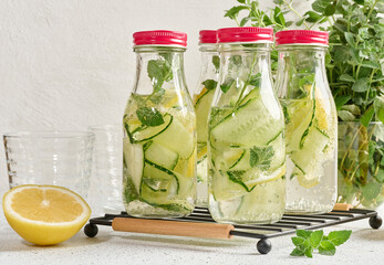 Glass bottles with red lids, filled with detox water containing cucumber and lemon slices and mint...