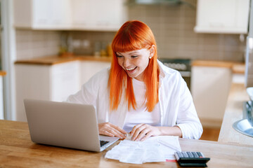 Bright-haired woman works on laptop at home, engaged in task with papers and a calculator on wooden table in modern kitchen