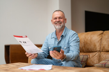 Man sitting comfortably on a sofa while reviewing documents and using a smartphone in a modern living room during the afternoon
