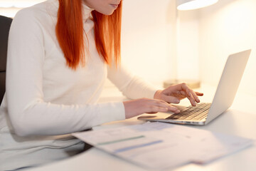 Fototapeta premium Focused woman working on a laptop at home during evening hours with paperwork on the desk and soft lighting