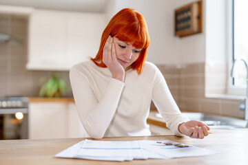 Young woman with red hair reviews documents at a kitchen table during a sunny afternoon in a bright...
