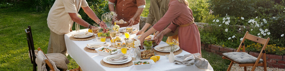 Family members arranging food and drinks on outdoor dining table in garden, hands reaching for...