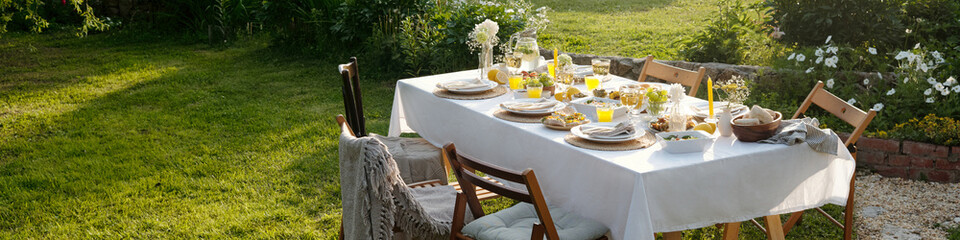 Outdoor dining table set for meal with plates, glasses, bread, salads, juice, flowers arranged on tablecloth, wooden chairs surrounding table in garden with green grass