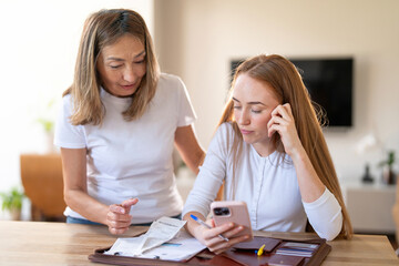 Two women discussing finances at home in a modern living space during daytime with papers and phone in hand