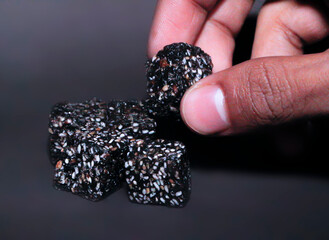 A Detailed Close-up of a Human Hand Delicately Holding a Single, Dark, Rustic Cube, a Healthy and Natural Snack Made from a Rich, Textured Paste and Generously Embedded with White Sesame Seeds