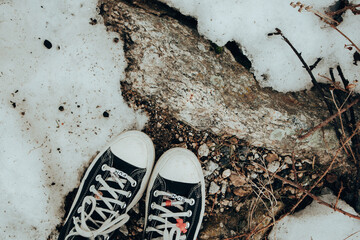Black canvas sneakers on snow and rock, fun-themed shoelace charms, in a top view. A pair of black...