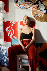 Vertical medium long portrait of young professional artist sitting relaxed in studio looking away