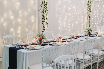 Elegant long wedding reception table with white chairs, candles, floral centerpieces and napkins tied with ribbons under soft lighting.