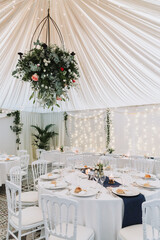 Floral chandelier with roses and greenery hanging over decorated wedding tables inside white draped tent with fairy lights.