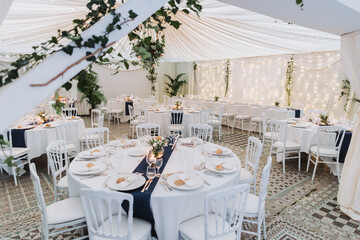 Wedding reception hall with round tables, white chairs, floral arrangements and string lights under elegant draped tent decoration.