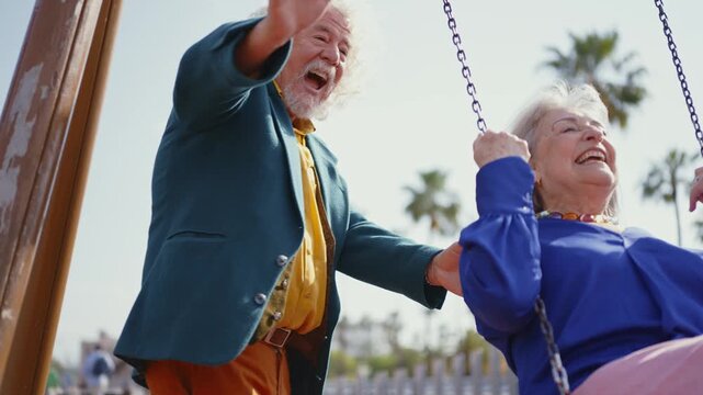 Elderly man pushing his wife on a swing set in a playground, both are laughing and enjoying their time together, showing their love and happiness