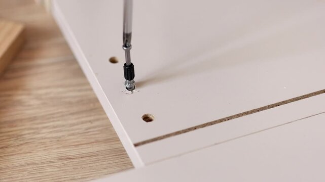 An overhead close-up shot of a screwdriver twisting a screw into a piece of flat-pack furniture.