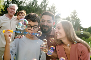 Multiethnic family group including senior Caucasian couple, young adult Caucasian woman, young Middle Eastern man, and preteen boy blowing soap bubbles outdoors, all smiling together