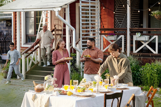 Group of multiethnic adults and one teenage boy gathering outdoors, smiling and preparing food around table set for meal, senior Caucasian man walking down steps in background
