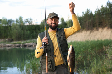 Smiling fisherman holding rod and caught fish near lake at summer