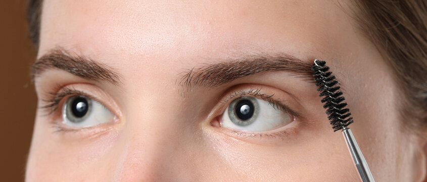 Young woman brushing her eyebrow with spoolie on brown background, closeup