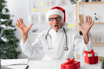 Elderly male doctor in Santa hat with shocked facial expression on workplace in the clinic....