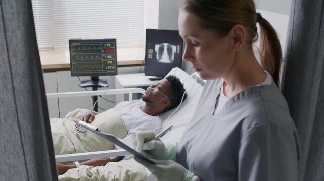 Young nurse taking notes on clipboard while checking on Black male patient with pneumonia, sleeping with oxygen tube and iv drip on hospital bed