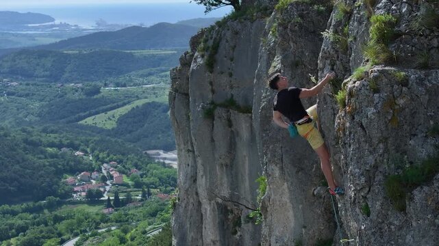 AERIAL: Young athletic man climbs a rock face and reaches the top hold of a climbing route. Male sport climber is training in a picturesque area surrounded by the Karst landscape above Adriatic Sea.