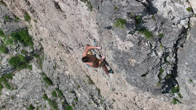 AERIAL, CLOSE UP: View from above of a shirtless male climber meticulously moving up a rock face. Young man tackles difficult climbing route on hot summer day. Adrenaline sport in natural environment.