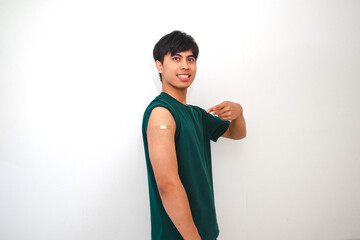 Smiling young Asian man showing arm bandage after vaccination, standing confidently on white background. Concept of vaccine, health care, and safe return to daily life