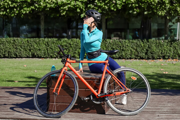 Thoughtful young woman in sporty outfit, wearing a helmet, talking on her phone while standing next...