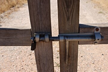 Close-up of a wooden fence gate with metal bolt and padlock in a rural field in Spain. Sunlit gravel track highlight traditional and the authentic character of andalusian landscapes