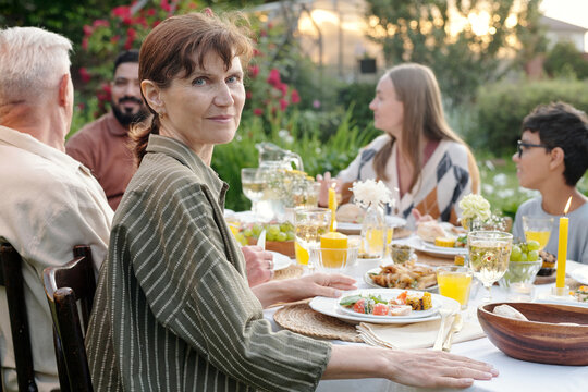 Portrait of middle aged Caucasian woman sitting at outdoor table with multiethnic family group enjoying meal together, woman looking into camera while others interacting