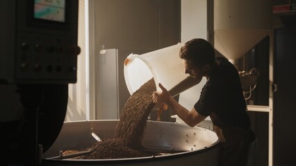 Caucasian male with short brown hair wearing brown apron while pouring roasted coffee beans from...