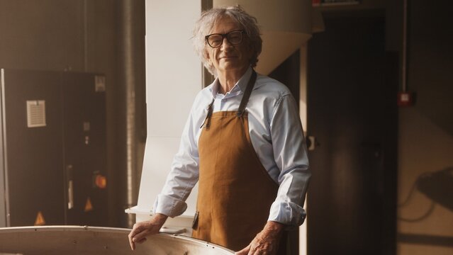 Confident Caucasian male with gray hair wearing glasses and brown apron while standing near coffee roasting equipment. Observing production process. Preparing to adjust settings for perfect roast.