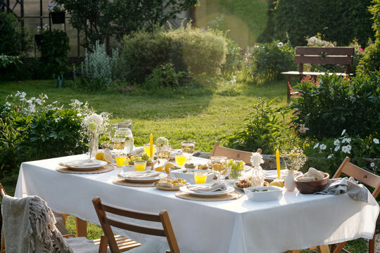 Outdoor garden table set for meal featuring plates, glasses of orange juice, bread, fruit, and flowers on tablecloth, wooden chairs surrounding table, lush greenery in background - Powered by Adobe