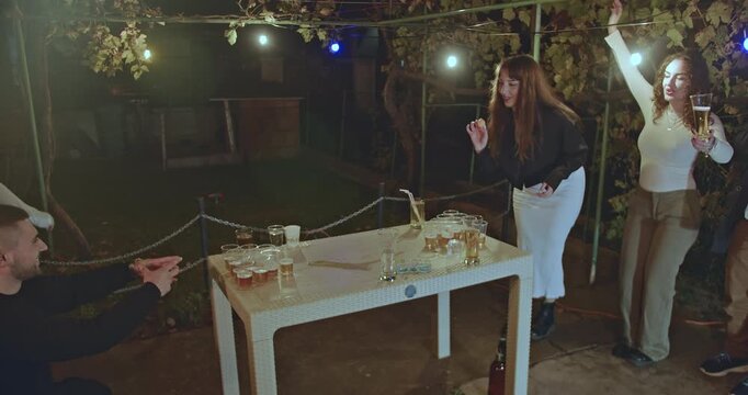 Friends playing beer pong in the backyard at night. A young man throws a ping pong ball, aiming at the cups filled with beer.