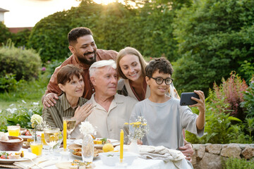 Multiethnic family group including senior Caucasian man, middle aged Caucasian woman, young adult Middle Eastern man, young adult Caucasian woman, preteen boy taking selfie outdoors at table