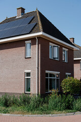 Brick house with solar panels on the roof, surrounded by greenery, under a bright blue sky.