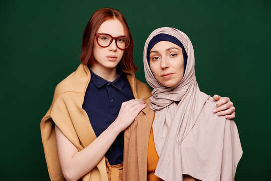 Horizontal medium portrait of modern young woman and her Muslim best friend standing together against tidewater green background looking at camera