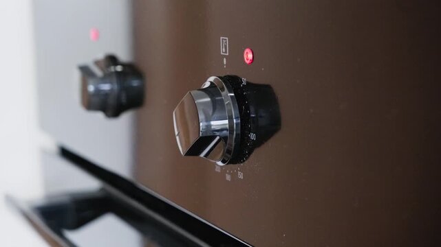 Hand operating dual oven control switches with red indicator lights. Close-up view of fingers turning knobs on modern appliance panel. Kitchen cooking preparation with clear tactile interaction.
