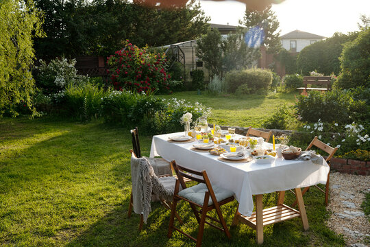 Outdoor table set for meal standing on green lawn in garden with plates, glasses, bread, fruit and flowers arranged for gathering, empty wooden chairs surrounding table