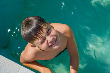 A young smiling boy is looking at the camera while standing in an outdoor swimming pool and holding...
