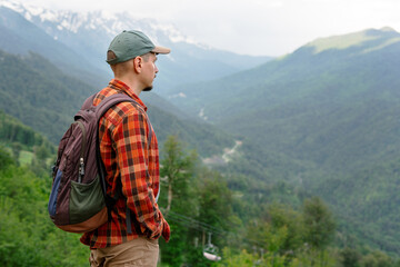 Male hiker wearing red plaid shirt , backpack and cap stands on a mountain overlook, gazing at the...