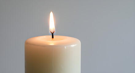 A lit candle emits a warm glow. The candle has a white wax body and is placed against a blurred grey backdrop