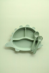 Mint green dinosaur-shaped kids plate with spoon and fork on soft pastel background. Minimalist baby tableware for playful and stylish mealtime moments.