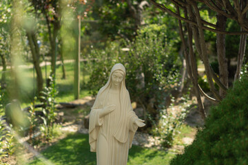 Fototapeta premium A white statue of the virgin mary is standing in a private lush green garden on a sunny day with blurred foliage in the background, concept of religion, spirituality, gardening