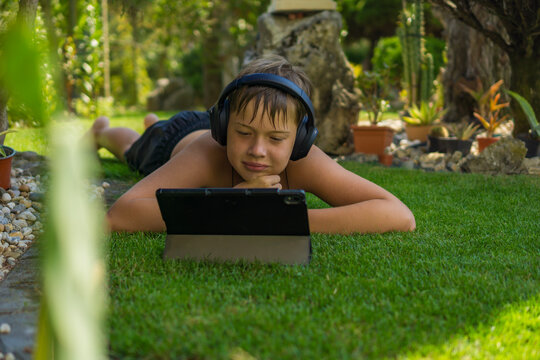 A boy is wearing headphones while lying on a lush green grass and using a tablet in a private garden on a sunny day, concept of family fun, children, technology