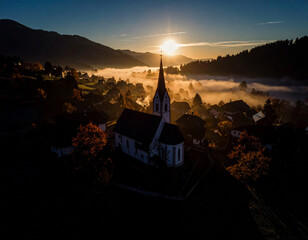 Misty November morning over small European village, church tower silhouette above fog, muted tones, cinematic aerial feel,